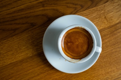 A top view of a small cup of espresso served on a white saucer, placed on a wooden surface. The espresso has a rich, dark brown crema on top.