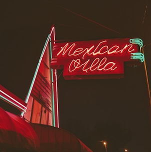A brightly illuminated neon sign in red and green advertises 'Mexican Villa' against a dark night sky. The sign features stylized text with a retro aesthetic and is attached to a building that appears slightly in the background.