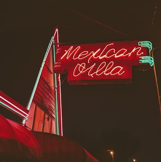 A brightly illuminated neon sign in red and green advertises 'Mexican Villa' against a dark night sky. The sign features stylized text with a retro aesthetic and is attached to a building that appears slightly in the background.