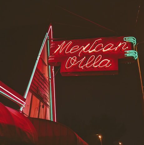 A brightly illuminated neon sign in red and green advertises 'Mexican Villa' against a dark night sky. The sign features stylized text with a retro aesthetic and is attached to a building that appears slightly in the background.