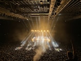 A large indoor concert venue is filled with a massive crowd, illuminated by bright stage lights beaming in multiple directions. The stage is set at the far end, with performers likely present, and the overhead lighting grid adds to the vibrant atmosphere. A visible haze from smoke or fog adds to the dramatic lighting effect.
