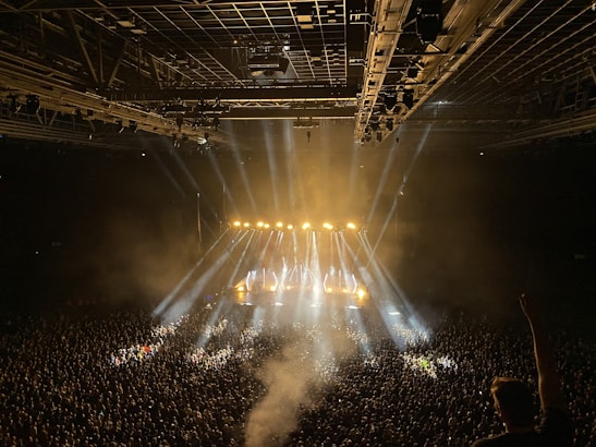 A large indoor concert venue is filled with a massive crowd, illuminated by bright stage lights beaming in multiple directions. The stage is set at the far end, with performers likely present, and the overhead lighting grid adds to the vibrant atmosphere. A visible haze from smoke or fog adds to the dramatic lighting effect.