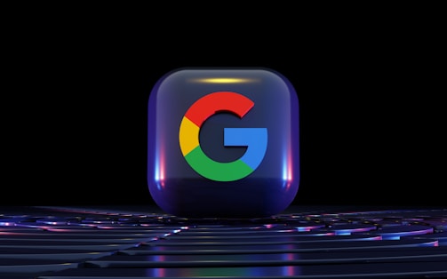 A three-dimensional glossy cube featuring the colorful Google logo against a dark background. The cube displays a reflective surface, capturing hints of surrounding lights, and is placed on a surface with subtle lighting effects.