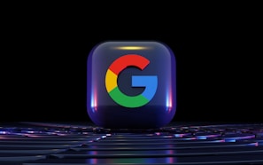 A three-dimensional glossy cube featuring the colorful Google logo against a dark background. The cube displays a reflective surface, capturing hints of surrounding lights, and is placed on a surface with subtle lighting effects.