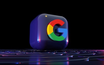 A 3D rendered cube featuring the Google logo prominently in the center. The logo is colored with its distinctive bright red, blue, yellow, and green. The cube rests on a surface with subtle light reflections, and the background is dark, creating a striking contrast.