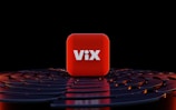 A three-dimensional red cube with the word 'ViX' in bold white letters positioned at its center, surrounded by a series of concentric, circular dark blue or black surfaces with orange highlights, set against a black background.