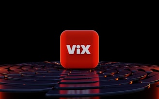 A three-dimensional red cube with the word 'ViX' in bold white letters positioned at its center, surrounded by a series of concentric, circular dark blue or black surfaces with orange highlights, set against a black background.