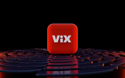 A three-dimensional red cube with the word 'ViX' in bold white letters positioned at its center, surrounded by a series of concentric, circular dark blue or black surfaces with orange highlights, set against a black background.