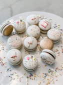 A dozen decorative macarons placed on a white plate, with festive and colorful sprinkles scattered around. The macarons are adorned with luxurious designs, including gold accents, small cakes, and balloon motifs.