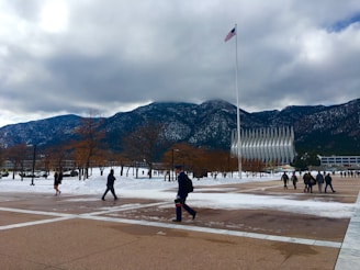 A snow-covered landscape with a large modern chapel featuring pointed spires set against a backdrop of mountains. People are walking on a plaza with scattered trees, and an American flag flies on a tall pole under a cloudy sky.