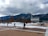 A snow-covered landscape with a large modern chapel featuring pointed spires set against a backdrop of mountains. People are walking on a plaza with scattered trees, and an American flag flies on a tall pole under a cloudy sky.