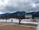 A snow-covered landscape with a large modern chapel featuring pointed spires set against a backdrop of mountains. People are walking on a plaza with scattered trees, and an American flag flies on a tall pole under a cloudy sky.