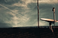 A satellite dish is positioned on a rooftop with a cloudy sky in the background. A contrail from an airplane is visible cutting diagonally across the sky. The foreground includes the silhouette of the rooftop and part of the building structure.