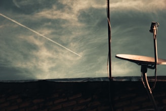 A satellite dish is positioned on a rooftop with a cloudy sky in the background. A contrail from an airplane is visible cutting diagonally across the sky. The foreground includes the silhouette of the rooftop and part of the building structure.
