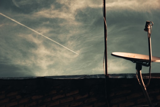 A satellite dish is positioned on a rooftop with a cloudy sky in the background. A contrail from an airplane is visible cutting diagonally across the sky. The foreground includes the silhouette of the rooftop and part of the building structure.