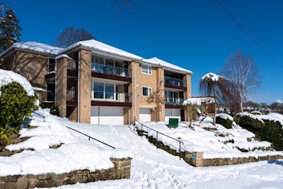 A multi-story brick residential building surrounded by a snowy landscape. The structure features several balconies with glass railings and white garage doors. Snow covers the sloping ground, stairs, and surrounding trees and bushes, indicating a winter setting. The sky is clear and blue, adding contrast to the scene.