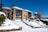 A multi-story brick residential building surrounded by a snowy landscape. The structure features several balconies with glass railings and white garage doors. Snow covers the sloping ground, stairs, and surrounding trees and bushes, indicating a winter setting. The sky is clear and blue, adding contrast to the scene.