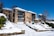 A multi-story brick residential building surrounded by a snowy landscape. The structure features several balconies with glass railings and white garage doors. Snow covers the sloping ground, stairs, and surrounding trees and bushes, indicating a winter setting. The sky is clear and blue, adding contrast to the scene.