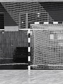 A black and white indoor sports hall with a focus on a futsal or handball goal, along with a net and wooden flooring. There are overhead lights visible in the background, and the scene is devoid of people.