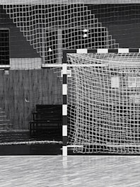 A black and white indoor sports hall with a focus on a futsal or handball goal, along with a net and wooden flooring. There are overhead lights visible in the background, and the scene is devoid of people.