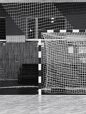 A black and white indoor sports hall with a focus on a futsal or handball goal, along with a net and wooden flooring. There are overhead lights visible in the background, and the scene is devoid of people.