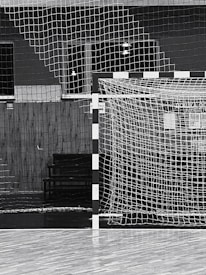A black and white indoor sports hall with a focus on a futsal or handball goal, along with a net and wooden flooring. There are overhead lights visible in the background, and the scene is devoid of people.