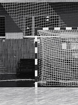 A black and white indoor sports hall with a focus on a futsal or handball goal, along with a net and wooden flooring. There are overhead lights visible in the background, and the scene is devoid of people.