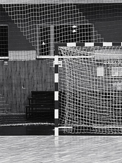 A black and white indoor sports hall with a focus on a futsal or handball goal, along with a net and wooden flooring. There are overhead lights visible in the background, and the scene is devoid of people.