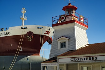 A large ship is positioned near a traditional lighthouse structure with a red and white color scheme. The ship's hull is prominently visible, with ropes attached to it. The building has a sign labeled 'CROISI&Egrave;RES' and is constructed with wooden siding and a small dormer window. The sky is clear and blue, indicating a sunny day.
