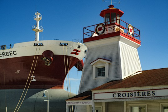 A large ship is positioned near a traditional lighthouse structure with a red and white color scheme. The ship's hull is prominently visible, with ropes attached to it. The building has a sign labeled 'CROISI&Egrave;RES' and is constructed with wooden siding and a small dormer window. The sky is clear and blue, indicating a sunny day.