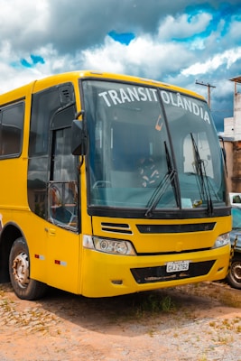 A yellow bus with the word 'TRANSITOLÂNDIA' displayed on the front is parked on a dirt surface. The bus has a distinct front design with large windows and visible windshield wipers. Part of a car is visible next to it. In the background, there are utility poles and buildings under a cloudy sky with patches of blue.