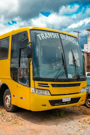A yellow bus with the word 'TRANSITOL&Acirc;NDIA' displayed on the front is parked on a dirt surface. The bus has a distinct front design with large windows and visible windshield wipers. Part of a car is visible next to it. In the background, there are utility poles and buildings under a cloudy sky with patches of blue.