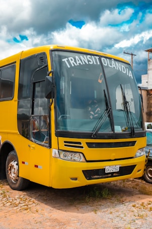 A yellow bus with the word 'TRANSITOL&Acirc;NDIA' displayed on the front is parked on a dirt surface. The bus has a distinct front design with large windows and visible windshield wipers. Part of a car is visible next to it. In the background, there are utility poles and buildings under a cloudy sky with patches of blue.