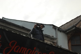 A person wearing a dark jacket is standing on a rooftop holding a smartphone and appears to be taking a photo or video. The building has a sign with 'Jamaican' written on it, and the sky is overcast.