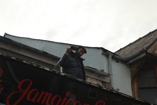 A person wearing a dark jacket is standing on a rooftop holding a smartphone and appears to be taking a photo or video. The building has a sign with 'Jamaican' written on it, and the sky is overcast.
