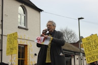 An older man is speaking passionately into a microphone at an outdoor event. He is holding a paper with the words 'STOP ZONING' on it. Around him are yellow placards with messages such as 'KEEP OXFORD FREE, NO TO 15 MINUTE CITY' and 'NO TRAFFIC FILTERS, DON'T DIVIDE OXFORD'. The background features a white brick building with a window and a yellow door. It is an overcast day.