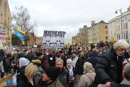 A crowd of people gathered in a public space, holding various signs and banners. There are signs with slogans related to media and social issues. The setting appears to be an urban area with historical buildings in the background. Some attendees are bundled up in coats and hats, suggesting cool weather.