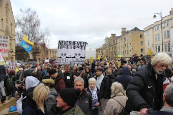 A crowd of people gathered in a public space, holding various signs and banners. There are signs with slogans related to media and social issues. The setting appears to be an urban area with historical buildings in the background. Some attendees are bundled up in coats and hats, suggesting cool weather.