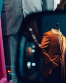 A storefront display features mannequins dressed in formal suits. A grey suit and an eye-catching orange blazer are visible. Reflections and bokeh lights add a dynamic and artistic flair to the scene.
