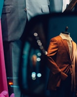 A storefront display features mannequins dressed in formal suits. A grey suit and an eye-catching orange blazer are visible. Reflections and bokeh lights add a dynamic and artistic flair to the scene.