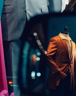 A storefront display features mannequins dressed in formal suits. A grey suit and an eye-catching orange blazer are visible. Reflections and bokeh lights add a dynamic and artistic flair to the scene.