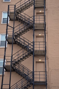 A multi-story building exterior with a metal fire escape featuring zigzag patterns is visible. The escape consists of multiple flights of black, metallic stairs with vertical railings, attached to a beige brick wall. Several closed doors and windows are evenly spaced along the staircase, and small light fixtures are mounted near each door.