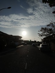 A residential street is depicted with the sun setting or rising in the background, casting a bright light. Houses line both sides of the street, with visible roofs and driveways. Cars are parked along the sides, and a streetlamp is visible silhouetted against the sky. Trees with sparse leaves are seen at the edges, and wispy clouds stretch across the blue sky.