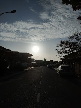 A residential street is depicted with the sun setting or rising in the background, casting a bright light. Houses line both sides of the street, with visible roofs and driveways. Cars are parked along the sides, and a streetlamp is visible silhouetted against the sky. Trees with sparse leaves are seen at the edges, and wispy clouds stretch across the blue sky.