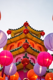 A vibrant, multi-tiered pagoda with colorful red, pink, and yellow lanterns hanging around it. The pagoda has ornate green roof edging and bright pink walls, creating a festive and lively atmosphere.