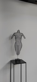 A wire sculpture of a human torso is displayed on a minimalist black stand. The sculpture has a hollow, mesh-like structure, showcasing the contours of a female torso without arms or a head. It is set against a plain, light-colored wall, creating a stark contrast.