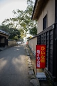 A narrow, serene pathway lined with traditional Japanese houses on the right side. A vibrant red sign with Japanese characters stands out near the entrance, adding a pop of color. The path is bordered by stones and shaded by trees, creating a calm, quiet atmosphere.