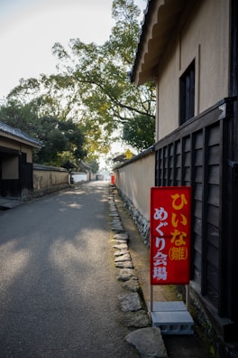 A narrow, serene pathway lined with traditional Japanese houses on the right side. A vibrant red sign with Japanese characters stands out near the entrance, adding a pop of color. The path is bordered by stones and shaded by trees, creating a calm, quiet atmosphere.
