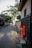 A narrow, serene pathway lined with traditional Japanese houses on the right side. A vibrant red sign with Japanese characters stands out near the entrance, adding a pop of color. The path is bordered by stones and shaded by trees, creating a calm, quiet atmosphere.