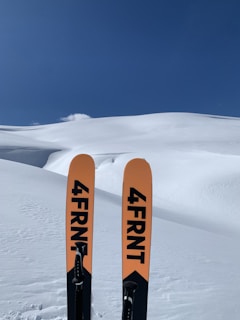 Two skis with the brand name '4FRNT' stand upright in the foreground against a backdrop of pristine, untouched snow and a clear blue sky.
